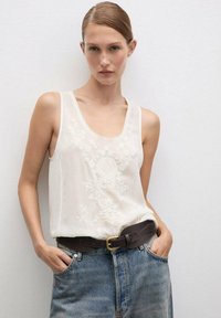Wit mouwloos topje met geborduurd bloempatroon, gecombineerd met high-waisted denim jeans en een bruine leren riem met een gouden gesp.