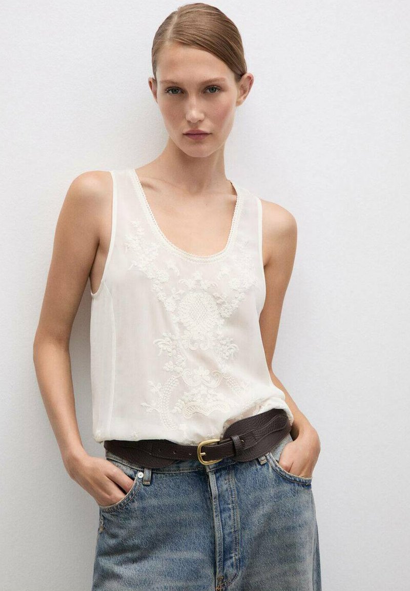 Wit mouwloos topje met geborduurd bloempatroon, gecombineerd met high-waisted denim jeans en een bruine leren riem met een gouden gesp.