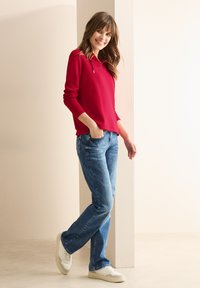 Haut à capuche rouge à manches longues, jeans droits bleus et baskets blanches. Le haut a une coupe décontractée et une capuche avec cordon. Design simple.