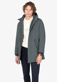 Joven con un abrigo de invierno gris con capucha sobre una sudadera de color claro y pantalones oscuros, de pie frente a un fondo blanco.