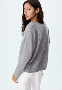 Ung kvinde med langt mørkt hår, iført en løs grå sweater og hvide bukser, vendt let til siden mod en ensfarvet lys baggrund.