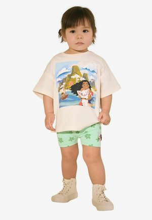Tricou din bumbac crem cu imprimeu grafic colorat, pantaloni scurți verzi cu model floral și adidași bej înalți. Ținută casual simplă.