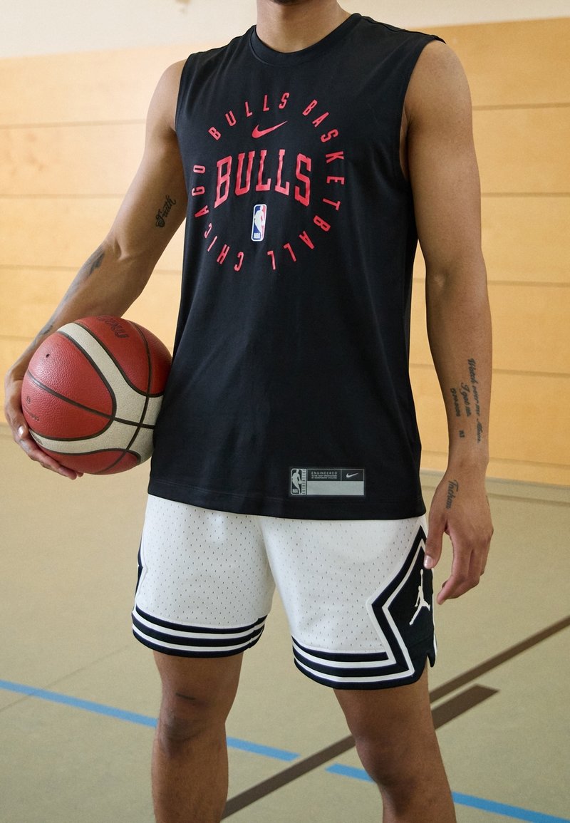Personne tenant un ballon de basket, portant un débardeur noir des Chicago Bulls et un short de basket blanc avec des bordures noires et le logo sur le terrain.