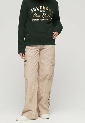 Osoba nosząca ciemnozieloną bluzę Superdry i beżowe spodnie cargo oraz białe trampki, stoi na jednolitym tle.