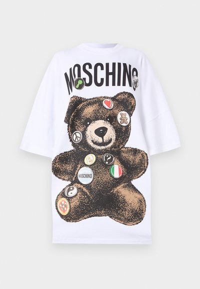 Bela oversize majica s kratkimi rokavi, ki prikazuje grafiko medveda okrašenega s pisanimi nalepkami in logotipom "Moschino" v debelih črnih črkah.