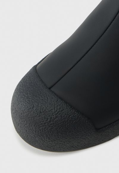 Prim-plan al vârfului unei pantofi negri, cu capac din cauciuc texturat și superior din piele netedă, cu cusături vizibile.