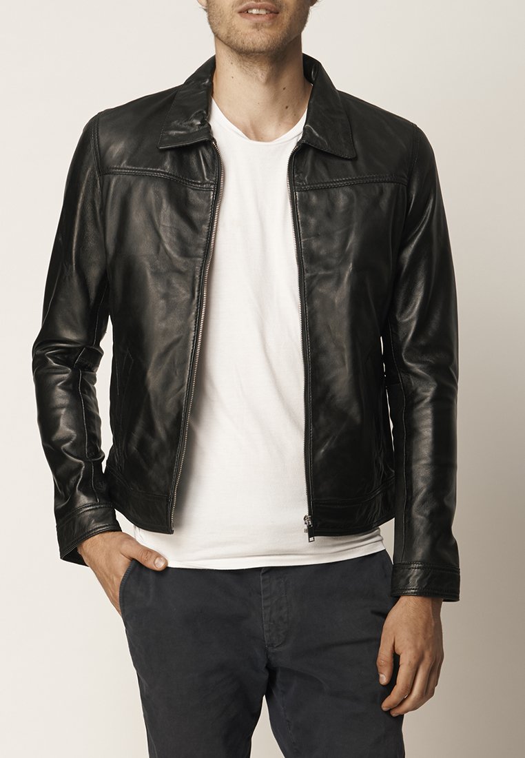 Giacca di pelle nera con chiusura a zip, colletto piatto e texture liscia. Presenta tasche laterali e una silhouette aderente. Indossata sopra una t-shirt bianca.