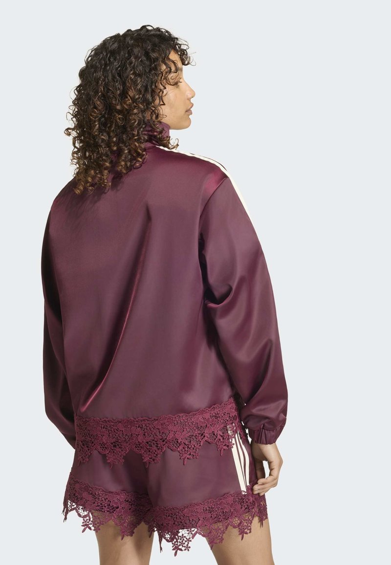 Femme aux cheveux bouclés portant une veste bordeaux brillante et un short assorti avec une bordure en dentelle florale, vue de dos sur un fond uni.