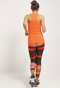 Un débardeur orange ajusté avec un dos nageur ; des leggings noirs avec des rayures horizontales rouge et orange. Chaussures de sport visibles.