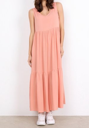 Perzikkleurige mouwloze maxi-jurk met een V-hals, geplooide taille en een gelaagde onderkant. Glad stof; gedragen met witte sneakers.