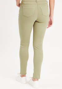 Jean skinny élastique vert olive avec taille haute, poches arrière cousues et longueur cheville, associés à des baskets blanches.