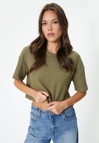 Camiseta corta de color verde oliva hecha de tejido texturizado, con mangas cortas y un único bolsillo en el pecho, combinada con jeans de color azul claro.
