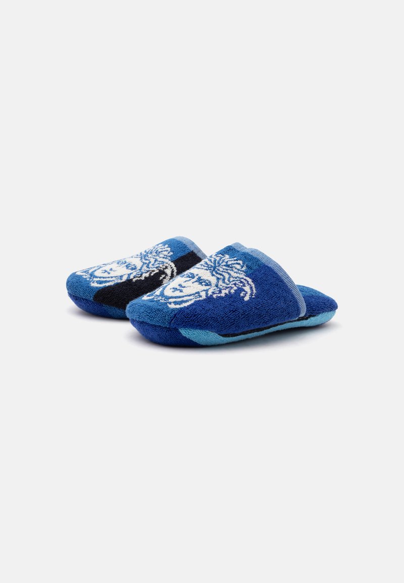 Paar blaue Slipper zum Hineinschlüpfen mit weißem dekorativem Gesichtsmotiv oben, auf einem schlichten hellen Hintergrund dargestellt.