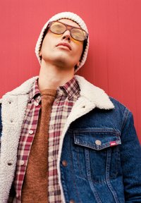 Denim jakk keskmise villase kraega, ruuduline särk punastes ja pruunides toonides, pruun kampsun all, ja kollakaspruunid prillid koos valge beanie'ga.