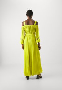Abito lungo a balze, scollato sulle spalle, di un vivace giallo neon. Presenta maniche lunghe e una vita cinchata con dettagli metallici decorativi.