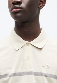 Μπεζ πόλο πουκάμισο από υφασμάτινη υφή με γιακά, που φέρει ανοιχτό γκρι ρίγα και κεντημένο λογότυπο "BOSS" στο στήθος.