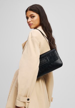 Borsa a tracolla in pelle nera con finitura testurizzata, dotata di placca con logo, indossata sulla spalla sopra un trench beige.
