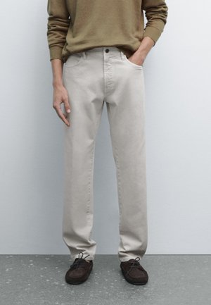 Person wearing beige pants, olive green sweater, and dark brown lace-up shoes standing against a plain light wall.