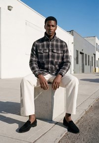 Homme portant une veste à carreaux noire, un pantalon blanc et des chaussures noires assis sur un bloc de béton sur un trottoir urbain ensoleillé.