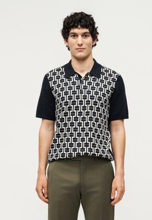 Fiatal férfi göndör hajjal, fekete-fehér mintás rövid ujjú galléros ingben és olívazöld nadrágban, egy egyszínű háttér előtt áll.