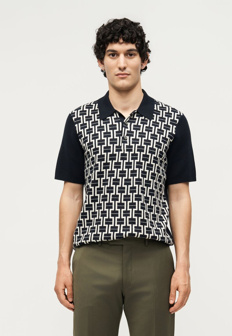 Νεαρός με σγουρά μαλλιά, φορώντας μαύρο-άσπρο πουκάμισο πόλο με κοντά μανίκια και ελιάς πράσινο παντελόνι, στέκεται μπροστά σε απλό φόντο.