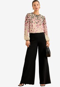 Blusa con stampa floreale e maniche trasparenti, che sfumano dal rosa alla crema. Pantaloni neri a gamba larga e una piccola borsa a tracolla nera texturizzata.