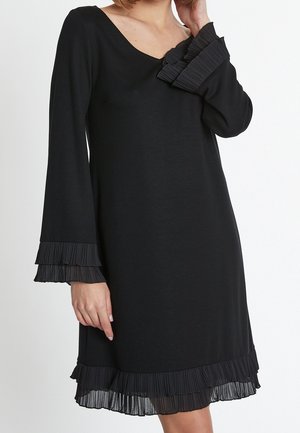 Femme portant une robe noire à manches longues avec des détails de volants plissés aux poignets, à l'encolure et à l'ourlet sur fond blanc.