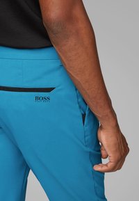 Modré sportovní kalhoty z texturované látky s černými akcenty na kapsách a logem na zádech. Pas je elastický pro pohodlí.