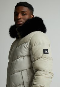 Puffjakke i lys beige med en svart faux pelskrage, kviltet design, og et logomerke på venstre erme. Glatt, matt tekstur.