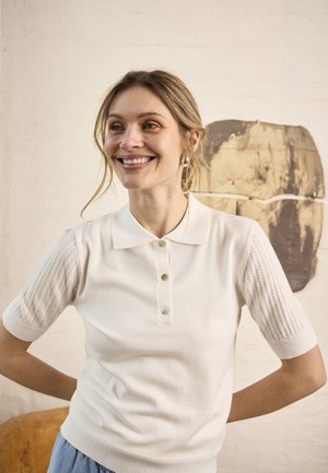 Femeie zâmbitoare cu păr blond purtând un tricou polo tricotat alb cu mâneci scurte, stând în interior, în fața unui perete deschis la culoare cu o lucrare de artă abstractă.