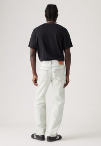 Białe dżinsy o luźnym kroju, widoczne tylne kieszenie i brązowa skórzana metka. Połączone z prostą czarną koszulą i czarnymi trampkami.