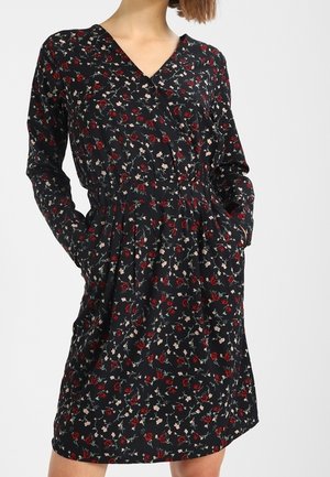 Vrouw draagt een zwarte lange mouwen jurk met kleine bloemenprints in rood en wit en heeft haar handen in de zijzakken.