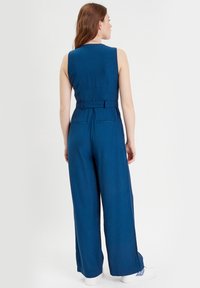 Tuta blu con design senza maniche, dotata di una vita con cintura, gambe larghe e due tasche posteriori. La texture sembra liscia e leggera.