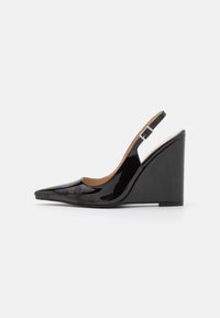 Scarpa con tacco a wedge in pelle verniciata nera, con punta affusolata e cinturino regolabile, mostrata in profilo laterale su sfondo bianco.