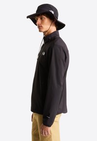 Μαύρο αδιάβροχο μπουφάν με ψηλό γιακά και τσέπες με φερμουάρ, συνδυασμένο με μαύρο καπέλο με φαρδύ γείσο, που παρουσιάζει έναν κομψό και λειτουργι κό σχεδιασμό.