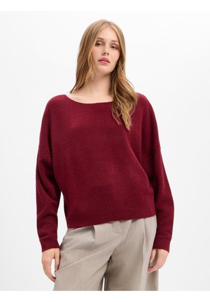 Jonge vrouw met lang blond haar, die een losse bordeauxrode trui en lichtbeige broek met wijde pijpen draagt, tegen een effen witte achtergrond.