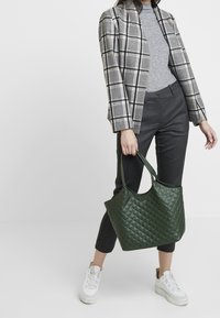 Gequilted groene leren tote bag met twee handvatten, gecombineerd met een grijze coltrui en een geruit blazer, zwarte krijtstreepbroek en witte sneakers.
