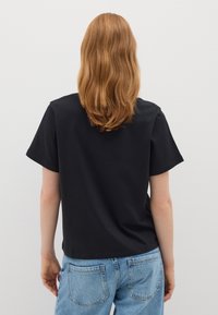 Persona con cabello largo y ondulado de color rojo, vistiendo una camiseta negra de manga corta sin estampado y jeans de color azul claro, de pie contra una pared blanca, de espaldas.