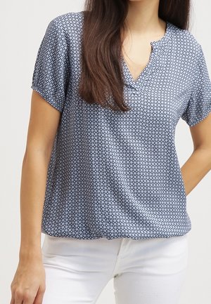 Femme portant un chemisier bleu à manches courtes avec un petit motif losange et un pantalon blanc, debout avec une main sur la hanche.