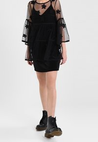 Czarna sukienka z warstwowym, przejrzystym nakryciem z aplikacjami kwiatowymi. Modelka ma na sobie czarne botki na sznurowanie.