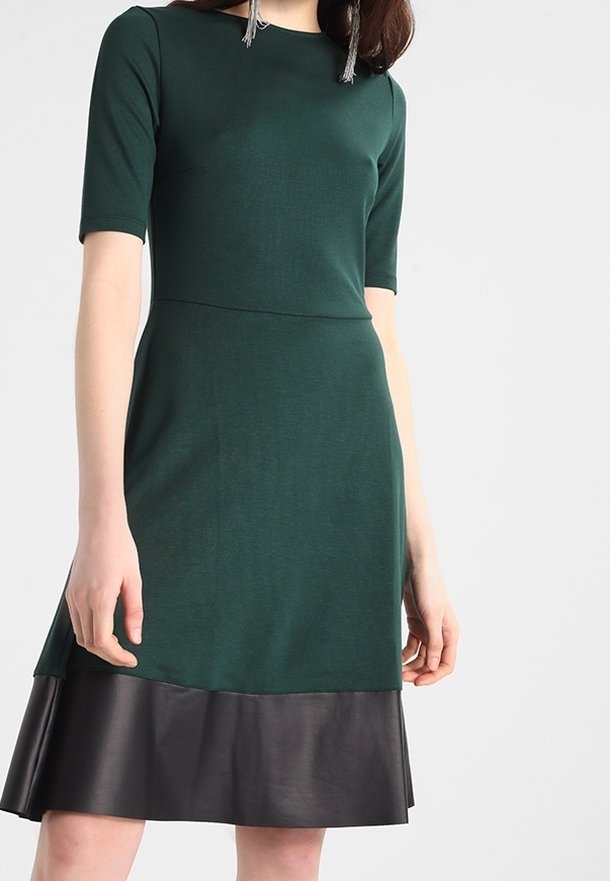 Femme portant une robe vert foncé avec des manches jusqu'au coude et une bordure en cuir noir à l'ourlet.
