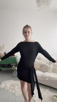 Donna in un abito nero asimmetrico con maniche lunghe, in piedi con le braccia distese in un luminoso soggiorno con un divano e un mobile verde.