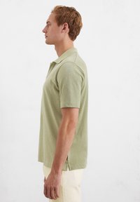 Polo vert à manches courtes avec col, doté d'ouvertures latérales pour plus de confort, fabriqué dans un tissu doux et texturé. Associé à un pantalon de couleur claire.