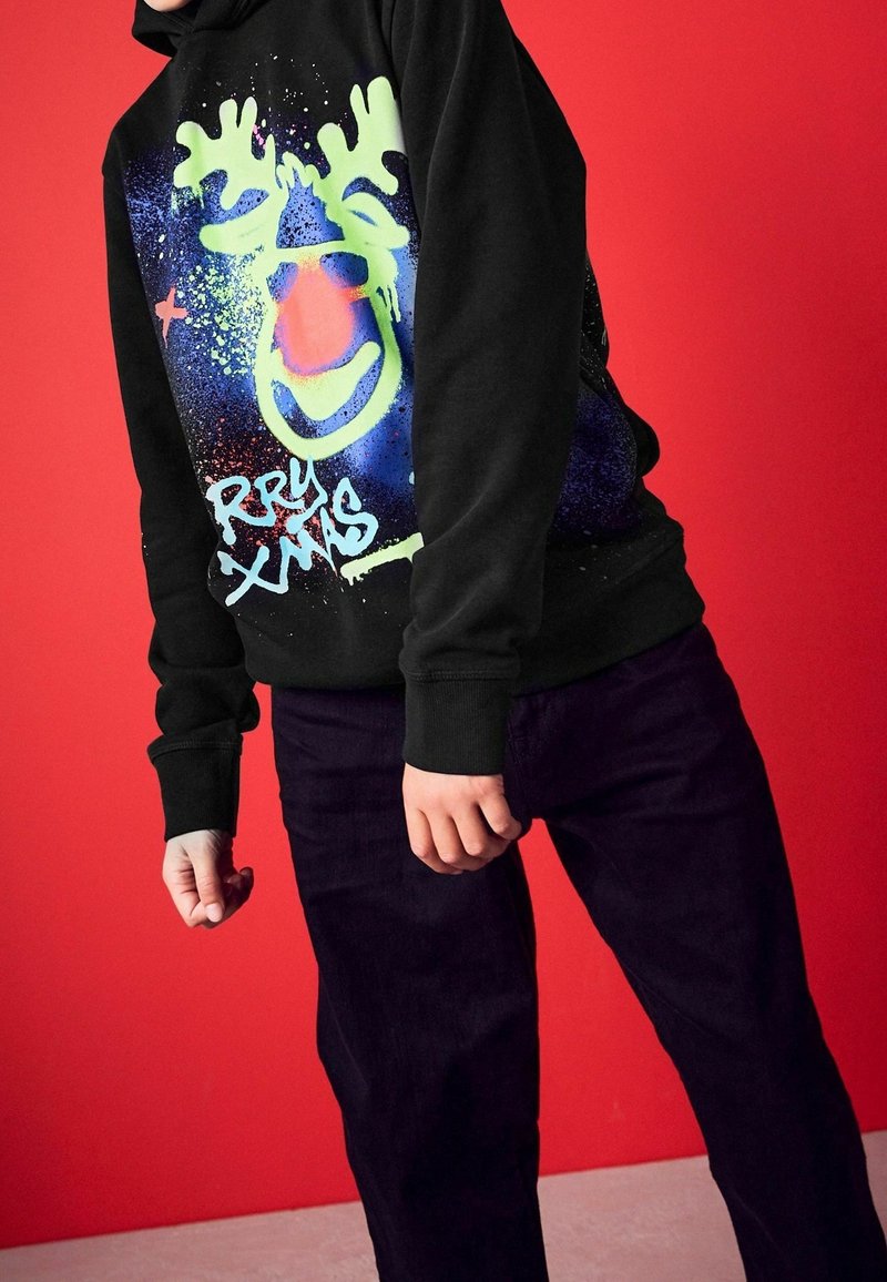Personne portant un sweat à capuche noir avec un visage de renne néon vert et le texte « RRY XMAS », associée à un pantalon foncé, sur un fond rouge.