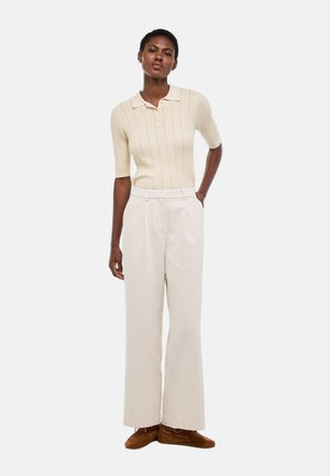 Femme debout portant un polo crème à manches courtes côtelé, un pantalon crème taille haute à jambes larges, et des mocassins en suède marron.