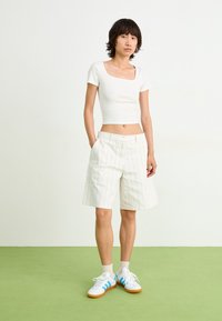 Top cropped branco com textura em canalé, calções de cintura alta em creme com riscas verticais finas e ténis brancos com detalhes azuis. Design simples e moderno.
