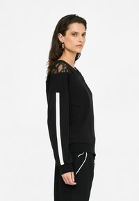 Sweatshirt noir avec détails en dentelle sur les épaules, accents blancs sur les manches et poignets côtelés. Le mannequin se tient de profil pour mettre en valeur la coupe et le design.