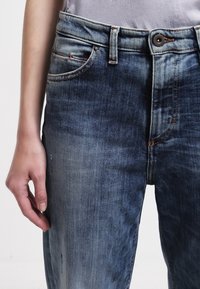 Närupplevelse av en persons hand som vilar bredvid urblekta blå denimjeans med synlig söm och knäppt midjeband.