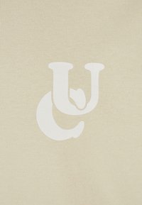 Krēmo krāsas audums ar teksturētu adījumu, centrā ir liels balts logotips, kas apvieno "C" un "U".