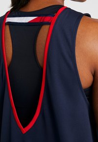 Marinblå sportstopp med djup V-bak, accentuerad av röda och vita band. Texturerad väv för ventilation. Lagerdesign som visar ett inre linne.